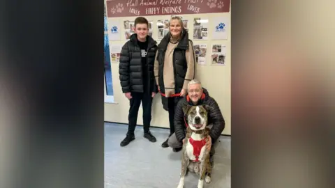 Hull Animal Welfare Trust A mother, father and teenage son, all wearing winter outdoor clothing, pose for a photo with rescue dog Winston. The father kneels behind the large dog with his hands resting on its sides. The mother and son stand behind. All are smiling.