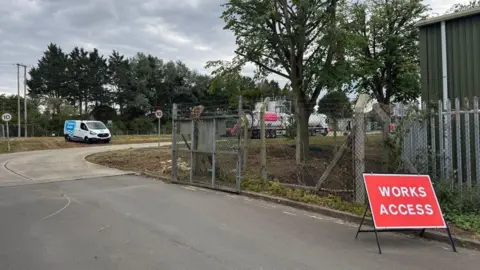 A view of the Wheatley Sewage Treatment Works site where a tank is parked and can be seen behind a metal fencing. There is a red sign for "Works access".