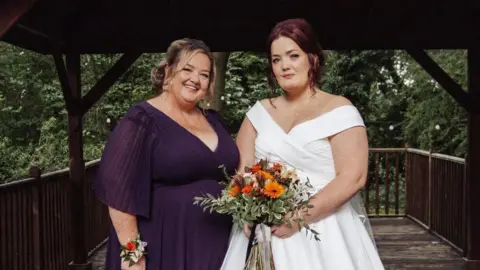 A woman was supplied to a woman in a purple dress, standing next to another woman in a white wedding dress, holding a bouquet of flowers. 