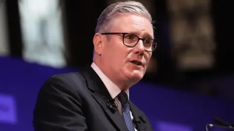 PA Media Sir Keir Starmer stands at a podium delivering a speech. He is wearing a dark suit, white shirt, and patterned tie, with short, combed hair and glasses. Behind him is a purple backdrop with blurred lights and indistinct text.
