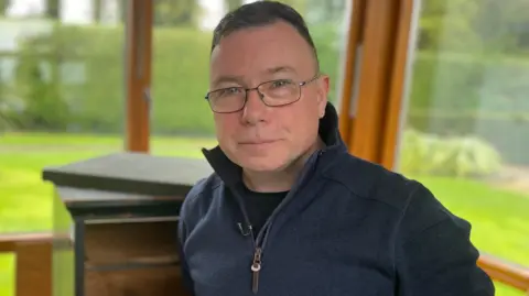 A man with short dark coloured hair is looking into the camera. He is wearing dark rimmed glasses, a navy jumper with a zip and a black t-shirt. The background is blurred, but he is standing inside with large windows behind him showing an open grassy area with trees.