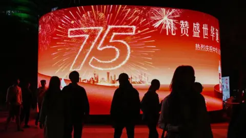 Getty Images La gente pasa junto a una pantalla gigante frente a un centro comercial que muestra un cartel que marca el 75.º aniversario de la fundación de la República Popular China, en el tercer día de una semana de vacaciones del Día Nacional en Beijing el 3 de octubre de 2024.