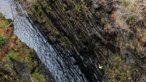 NTS A contractor wearing protective clothing and a hard hat abseils down into the gorge. The crag's rocks are dark and wet. Water flows at the bottom of the gorge.