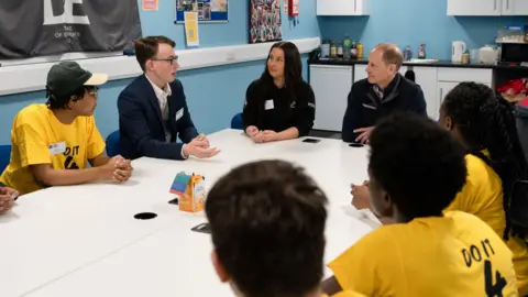 DofE Young people wearing T-shirts with around a table with the Duke of Edinburgh and two other adults, chatting.