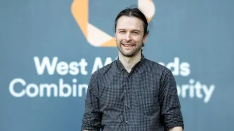 West Midlands Combined Authority A man with long dark hair tied back and dark facial stubble. He is wearing a navy and lighter blue checked shirt