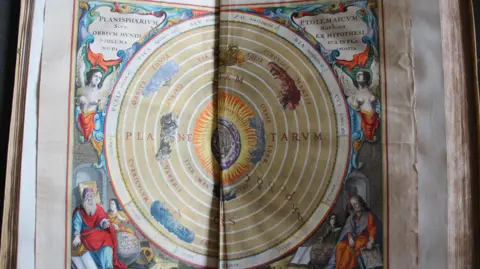 National Trust An elaborate and colourful double page illustration of planets in a circular pattern, with Latin descriptions and depictions of biblical and celestial figures in the corners. The pages are coming away in the middle and the edges are tattered.