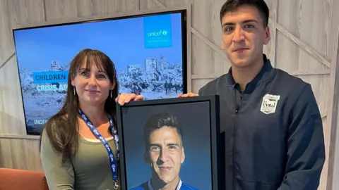 John Fairhall/BBC Louise Cobbold standing beside Marcelino Núñez, holding up her portrait of him between them. She has long dark brown hair with a fringe and is wearing a green long sleeved top. He has short dark hair and is wearing a dark grey zipped up jacket. They are standing in a room with a large screen behind them. 