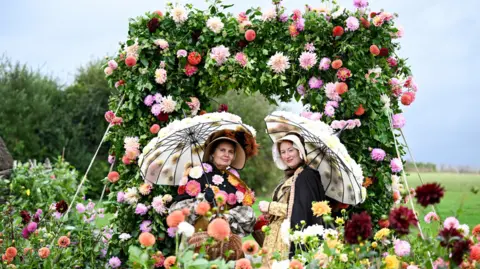 Getty Images Two ladies dressed in Victorian costume with parasols and surrounded by floral displays and an arch covered in trailing blossoms