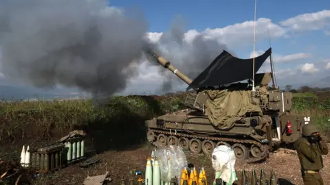 EPA An Israeli soldier stands in front of a self-propelled artillery howitzer in northern Israel as it fires a shell towards southern Lebanon (4 January 2024)