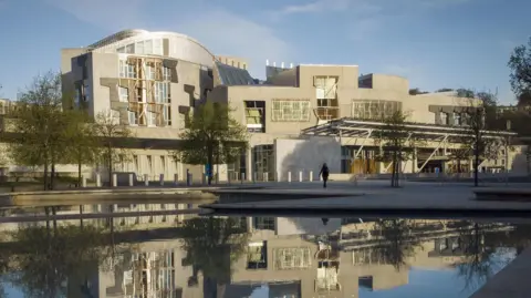 PA Media The Scottish Parliament building in Edinburgh with the building and trees reflecting on the water. 