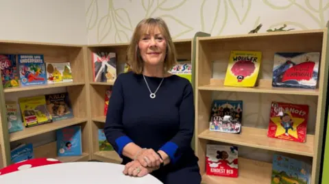 Staffordshire County Council A woman kneels in a school library at a table. She is wearing a navy dress with royal blue cuffs and a silver necklace with circular charm. She has brown hair with fringe. On the wooden shelves behind her are picture books.