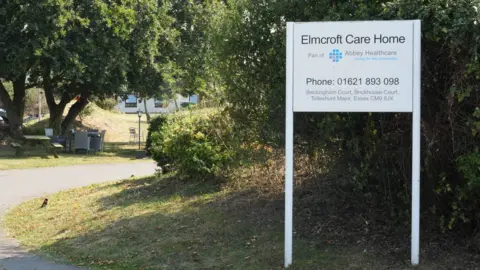 The entrance to Elmcroft Care Home, which has a white sign on the driveway. There are various trees and bushes next to the driveway.