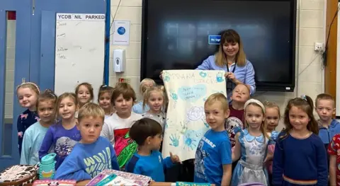 Wellingborough Polish School Lots of children with a teacher who is wearing a blue shirt and holding up a piece of paper