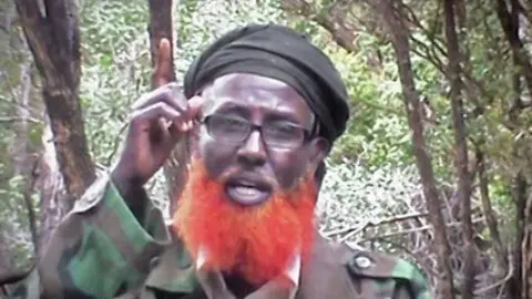 al-kataib Abdulqadir Mumin, with an orange beard and wearing reading glasses, fatigues and a black headscarf, hold ups his finger to the sky as he talks. Trees and bush can be seen behind him.