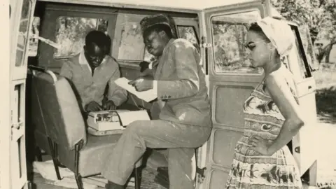 Herbert Weiss Pierre Molilli, on one foot inside the truck, looking at a piece of paper. A man inside the truck uses a typewriter, while André Blouin stands outside the truck with her hands on her hips.