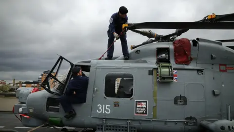 Two Royal Navy crew members in dark overalls work on a Wildcat/Lynx helicopter as it sits on the deck of a ship. The helicopter is dark grey and has the Royal Navy emblem on its side