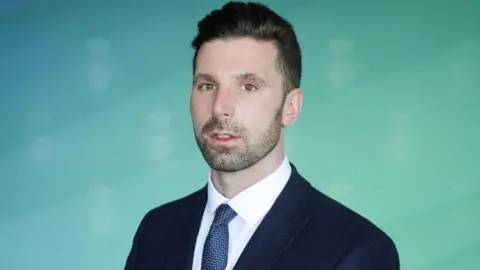 Merseyside Police Detective Sergeant Danny Gavine has short dark hair, a short beard, and is wearing a navy jacket, blue tie, and white shirt.
