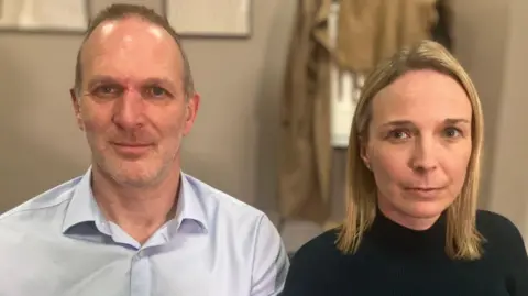 A landscape photograph of Jack and Sarah Hawkins sitting next to each other. Jack is on the left wearing a light blue shirt. Sarah is sitting on the right wearing a black roll-neck jumper.