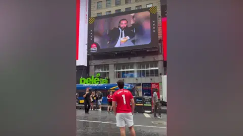Facebook A man is pictured wearing a red top, looking up at a screen in Times Square in New York which has a picture showing Jimmy Ward 
