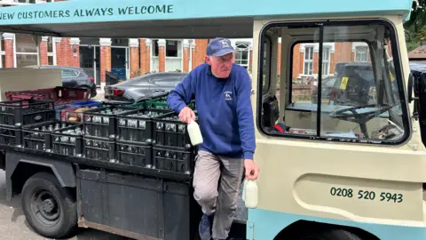 BBC/Harry Low Man in a blue Parker Dairies hat and jumper with cow logo climbs off a milk cart while holding three bottles of milk. There are black crates with milk bottles in the back of the cart. There are residential homes in the background. 