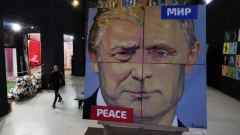 Reuters Giant painting showing composite face of Trump and Putin divided into quarters with the word peace in Russian and English at the top right and bottom left corners. It stands in a gallery in St Petersburg and the artist, Alexei Sergienko, is seen walking to the left of the picture.