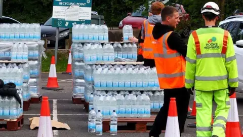 Several pallets of water bottles with road cones and a number of people in hi-vis outfits.
