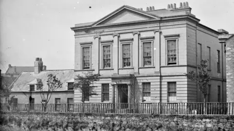 National Library of Ireland A black and white archive photo of Newry Savings Bank - a two-storey neoclassical building with a granite portico, five tall windows on its upper floor, separated by four decorative pilasters. There is a metal railing in front of the building which runs along a stone wall which skirts the river below. A small house with two dormer windows is sited to the left of the bank.