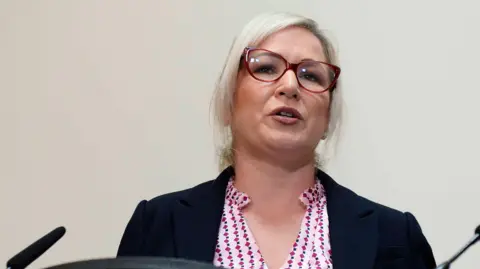 PA Michelle O'Neill mid speech with red glasses and pink shirt and navy blazer on a media platform