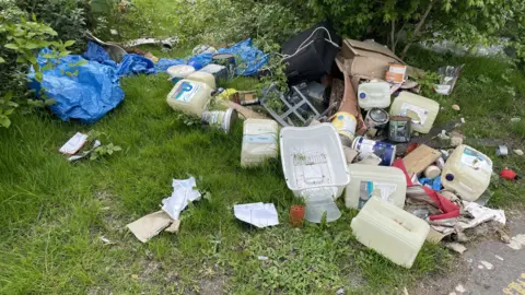 Plastic waste, including large bottles and tubs used for paint, are pictured abandoned on a grassy verge beside a road. There are blue sheets of plastic and cardboard among the rubbish, which lies beside some green bushes.