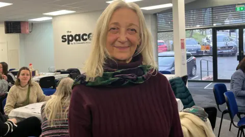 Anne Pierson-Smith with a burgundy jumper and a green scarf, standing in front of a room full of tables and chairs at St Oswalds Hospice Ouseburn building. 