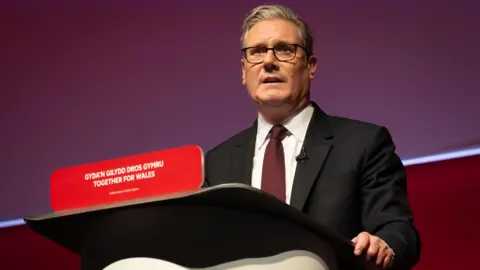Getty Images UK Prime Minister Sir Keir Starmer speaks during the 2025 Welsh Labour party conference on June 28, 2025 in Llandudno, Wales. He wears a dark suit, white shirt and maroon tie, and is standing behind a lectern with "Together for Wales" written in both English and Welsh.