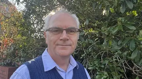 A man in a light blue shirt and navy vest wearing glasses is standing in front of green foliage and a brick wall looking to the camera
