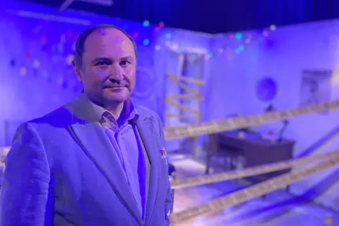 Jose stands in front of a display of a crime scene lit up in blue lights. The scene is a set of a bedroom or living room with a desk in it. The area is sealed off with police tape. 