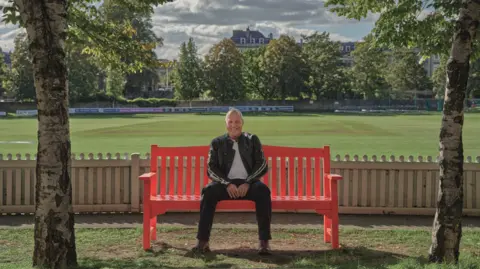 Bristish Heart Foundation Lee sitting on a red bench framed by two tree trunks in the foreground and with a cricket pitch behind him surrounded by mature trees. He is smiling and wearing a black track suit