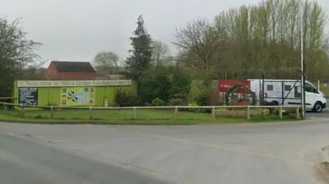 The entrance to Silica Lodge Garden Centre. There is a sign reading "Welcome to Silica Lodge Garden Centre". A white van is parked in the car park. The sky is overcast.