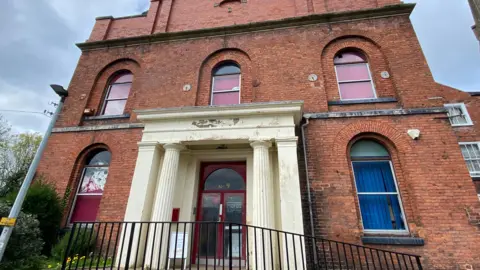 The front of a former chapel. It is red brick and has white painted pillars either side of the door. Its door is red and the windows have red blinds.