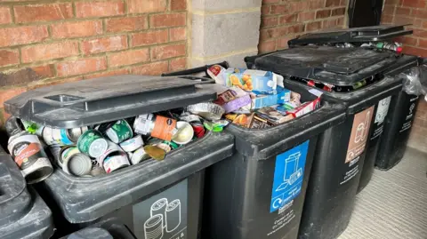 Five black bins lined up against the wall. They are full of recycling which means their lids are not fully closed. 