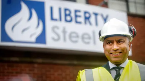PA Media A man in hi-vis and a helmet stands in front of a brick wall with a big sign attached. The blue and white sign reads "Liberty Steel" and features a logo on the left.