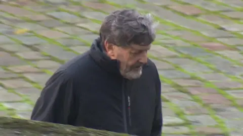 BBC Alan Harris walks towards Winchester Crown Court. He is wearing a black coat which has been zipped up. He has short greying dark hair and a greying beard.