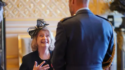 PA New Wendy Daunt standing in front of the Prince of Wales with smiling. She is wearing a navy fascinator in her hair and the image shows the back of Prince William who is facing her.