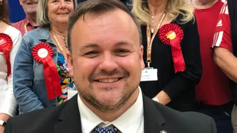 A smiling Terry Jermy looks directly at the camera in a suit, shirt and tie. He is standing at a count and is surrounded by others smiling and wearing red Labour rosettes. 