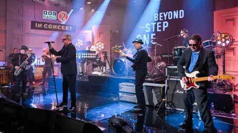 The band Madness perform on stage at a BBC Radio 2 concert. One man is holding a microphone while the others are playing various instruments.
