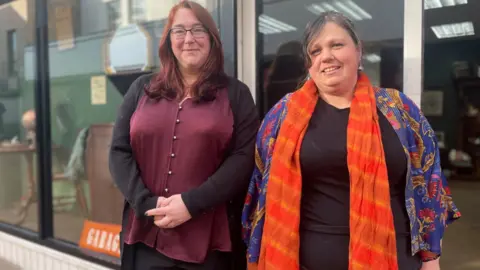 Simon Thake Two women in brightly coloured clothing stand together outside a shop front