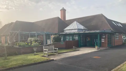 A single-storey hospice building viewed from outside the front entrance. A bench can be seen on the pathway leading to the door.