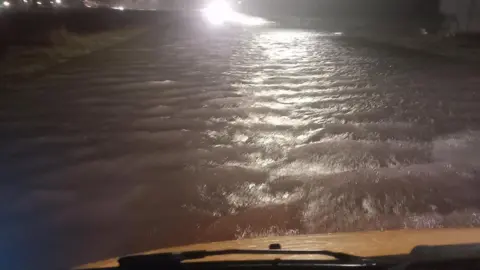 View of a flooded road at night taken from inside a vehicle. The white bonnet and windscreen wipers are visible at the bottom of the screen. Deep water lit up by street lights stretches out into the distance.