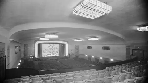 Historic England Archive A large curved cinema auditorium viewed from the back, with rows of wooden seats descending toward a stage. There are square lights on the ceiling and small lights on the walls.