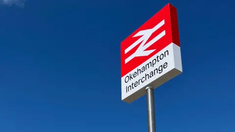 Alison Stephenson A sign with the words 'Okehampton Interchange' on it. It's red at the top and white below. The sky is blue.
