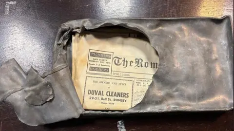 A large lead box is pictured with part of its front prised open. Inside is a newspaper, slightly faded, which is from 1969. 
