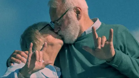 A man with white hair and a beard, wearing a green jumper, kissing a woman with short white hair, both are holding their hands up showcasing sign language