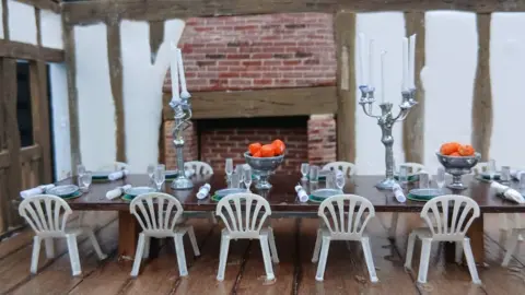 Simon Evans A model of the interior of a cottage where there is a long table that has glasses and plates placed on it. There are also white seats, chandeliers and bowls filled with oranges. There is a fireplace behind the table. 